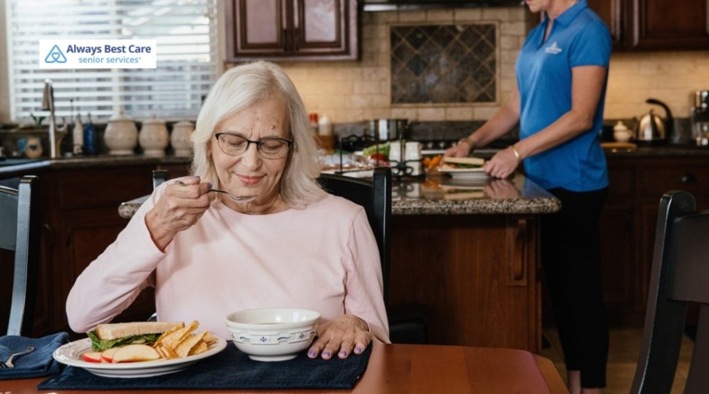 Senior woman eating soup at the kitchen table while a caregiver prepares a fresh sandwich behind her, illustrating mealtime assistance at home.