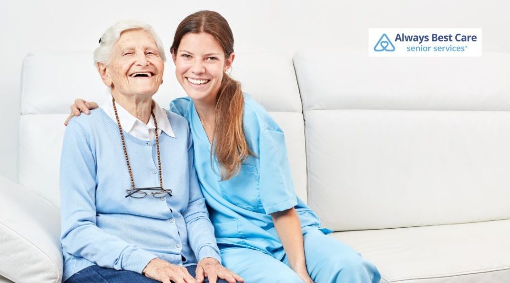 A joyful senior woman laughing alongside her caregiver, seated together on a white couch. The caregiver is offering her emotional support and companionship."