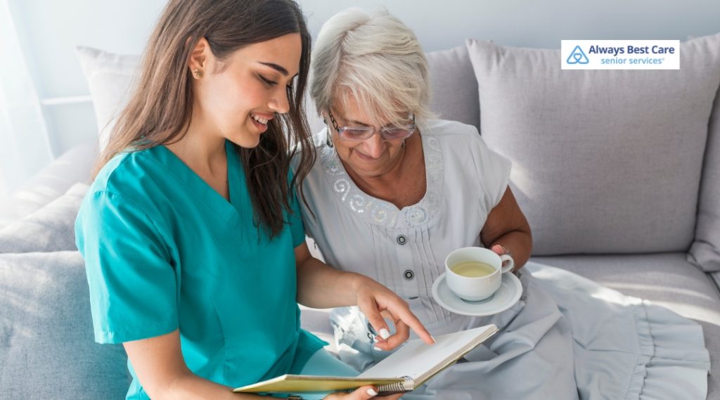 A caregiver and an elderly woman are sitting together on a sofa, looking at a book as the caregiver points at something, with the elderly woman holding a cup of tea.