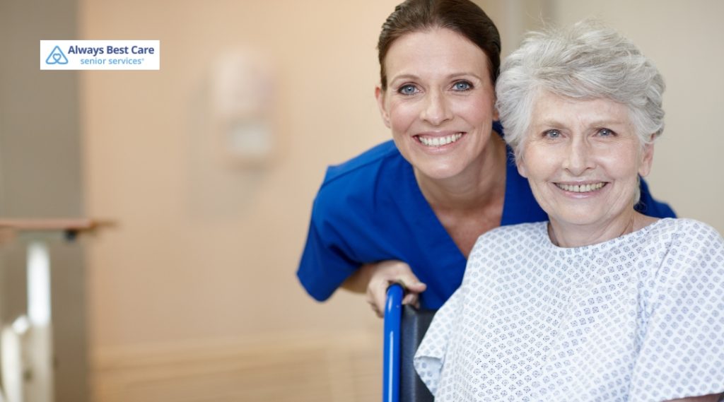 A caregiver and an elderly woman are smiling while the caregiver stands beside her in a wheelchair, showing kindness and support in a comfortable environment.