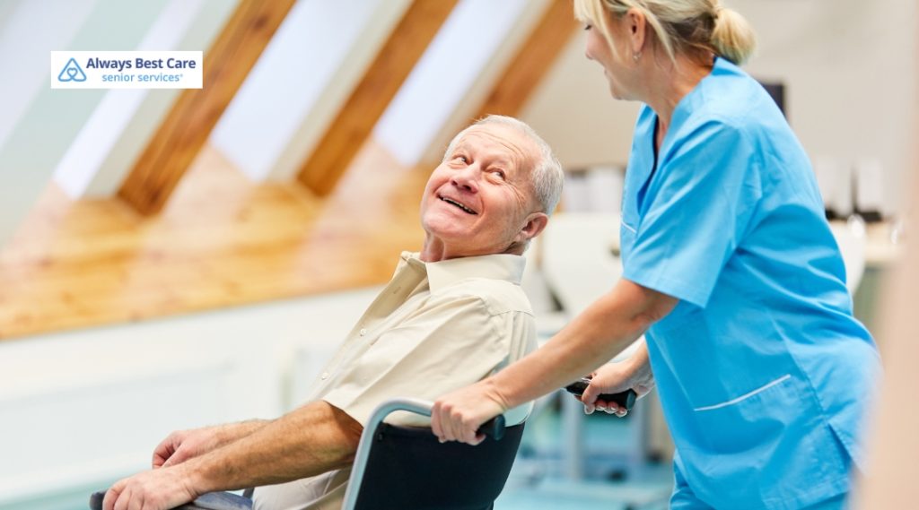 A caregiver is pushing an elderly man in a wheelchair while he smiles and looks at the caregiver in a bright and airy room.