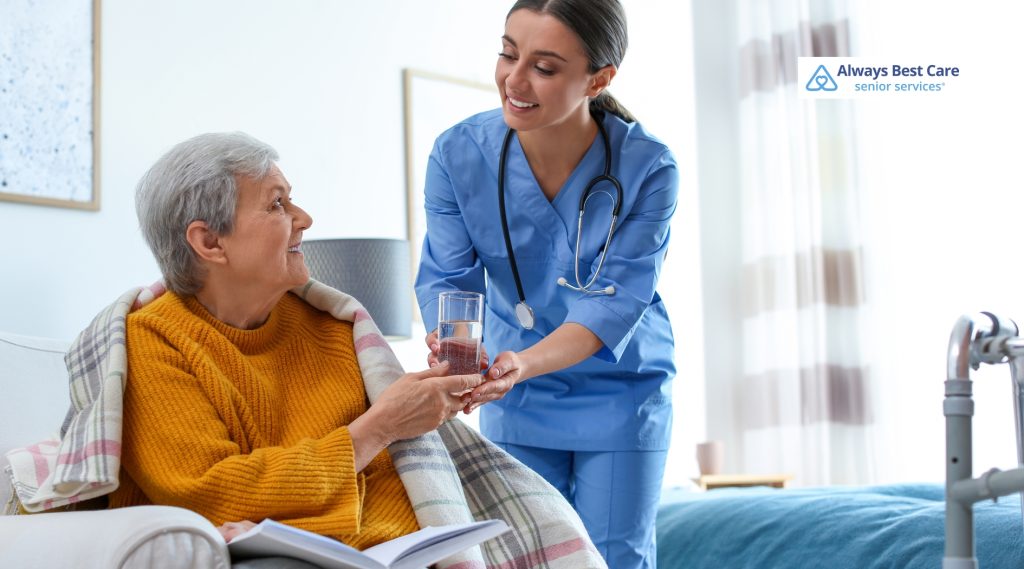 A caregiver handing a glass of water to a senior woman sitting on a couch, with the "Always Best Care" logo visible.