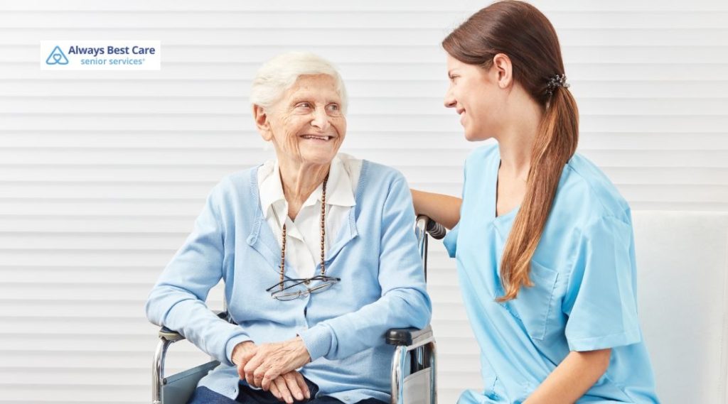 A senior woman in a wheelchair smiles brightly at her caregiver, who is seated beside her, both sharing a moment of warmth and connection.