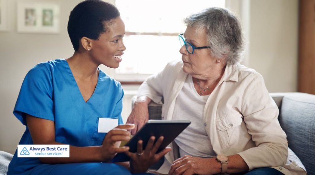 A caregiver and senior woman having a conversation while looking at a tablet. The caregiver is smiling, and the woman is listening attentively, with a comfortable home environment in the background.