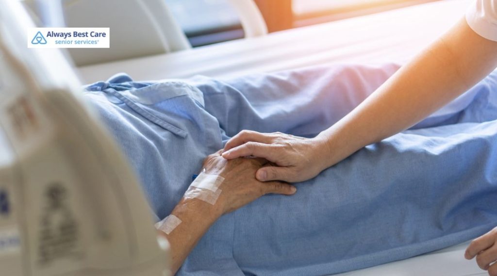 A compassionate caregiver holding a senior patient’s hand while they lie in a hospital bed. The patient is wearing a blue hospital gown, and the image conveys a sense of care and comfort.