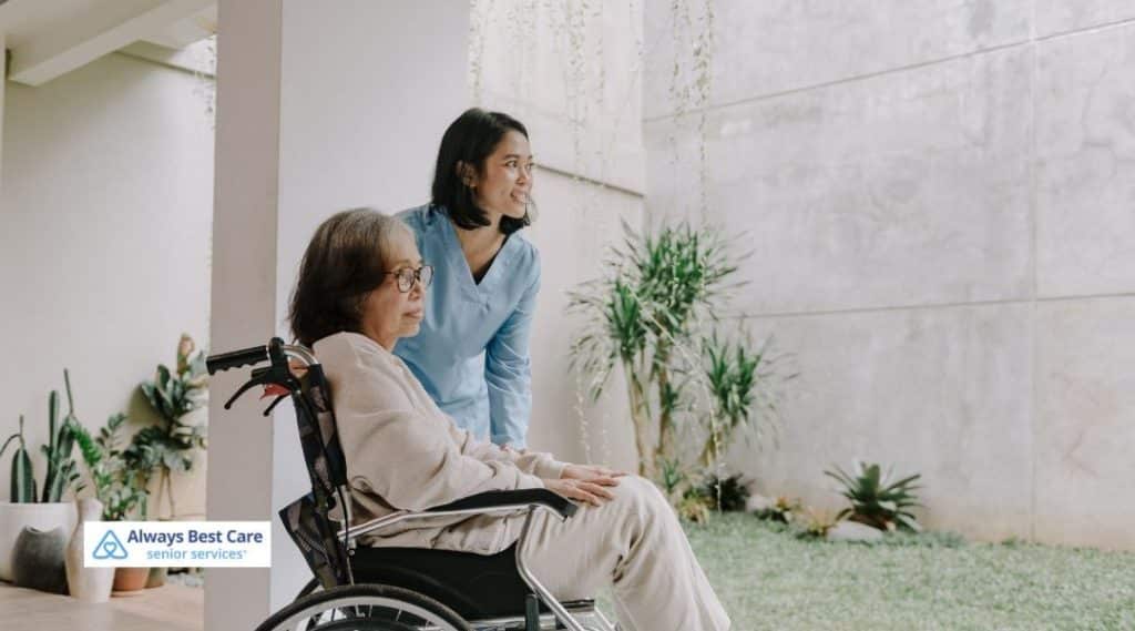 Senior woman in a wheelchair with a caregiver standing beside her, both enjoying the outdoors in a garden, with a sense of tranquility and care.