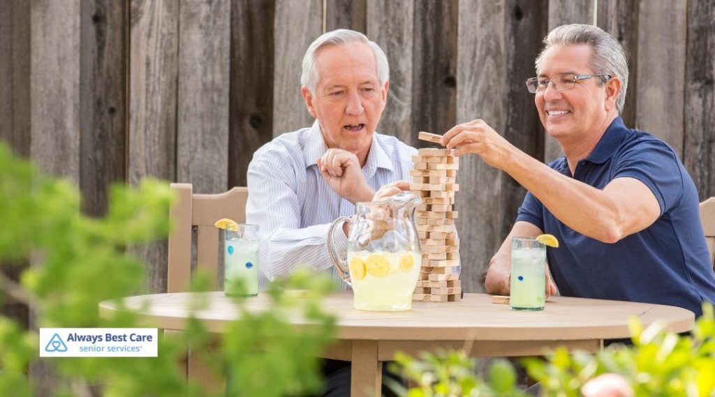 Group of smiling seniors sitting at a table stacking bright wooden blocks together during a memory care activity, Always Best Care senior services logo in the corner.
