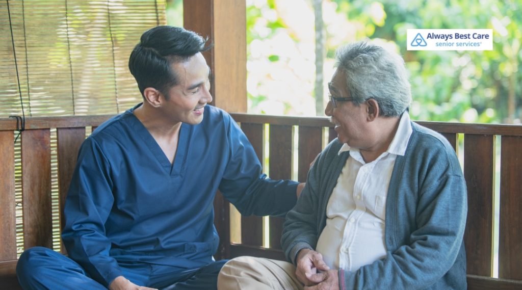 A caregiver in a blue uniform sits next to an elderly man in a relaxed outdoor setting, offering a comforting presence as they have a conversation. Both are smiling and seem to be enjoying each other's company.