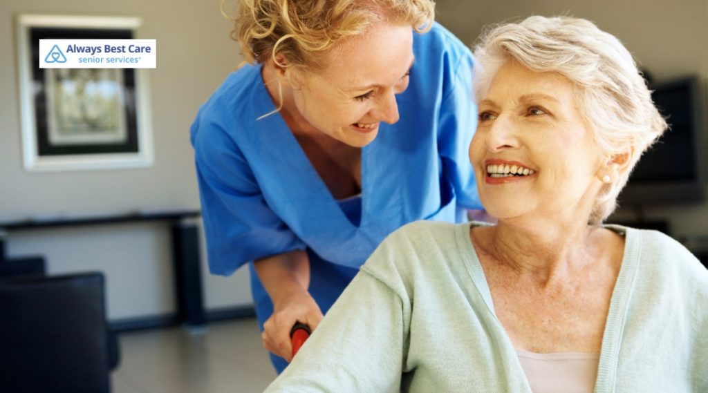 A caregiver, smiling and wearing blue scrubs, helps an elderly woman in a wheelchair. The elderly woman looks at the caregiver with a warm, genuine smile, both enjoying a moment of care and connection in a cozy indoor space.

