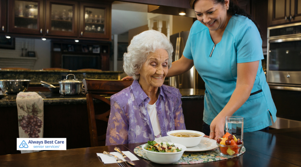 A caregiver in blue scrubs placing a bowl of soup in front of a senior woman wearing a purple floral blouse, sitting at a dining table, smiling in a cozy kitchen setting.