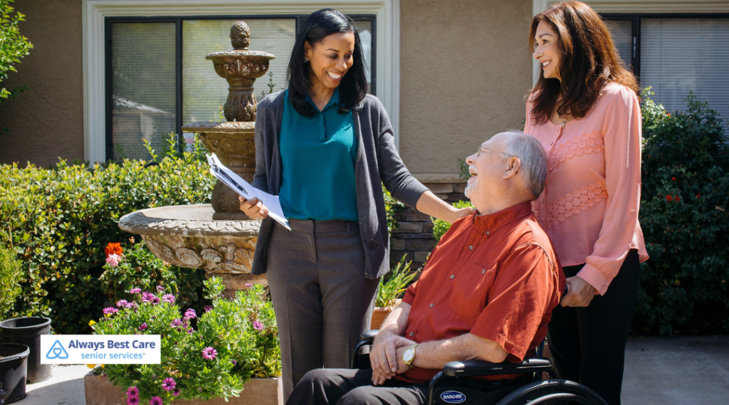A caregiver and two family members smiling and talking outside by a fountain, with the senior in a wheelchair, enjoying the warmth of family support. Always Best Care senior services logo visible at the bottom.
