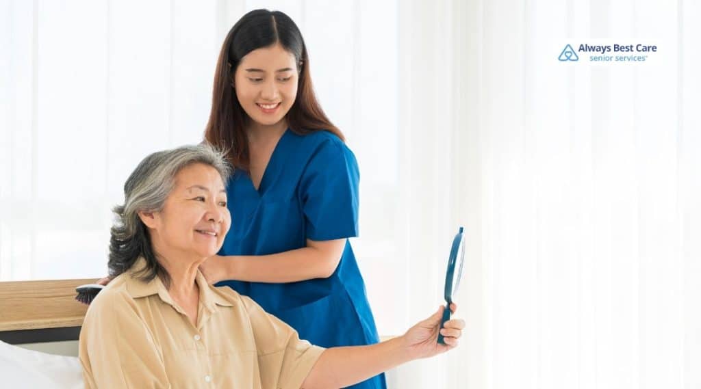 A caregiver in blue scrubs helps a senior woman sitting in a chair by handing her a mirror so she can see herself. Both women are smiling in a warm, comfortable indoor setting.