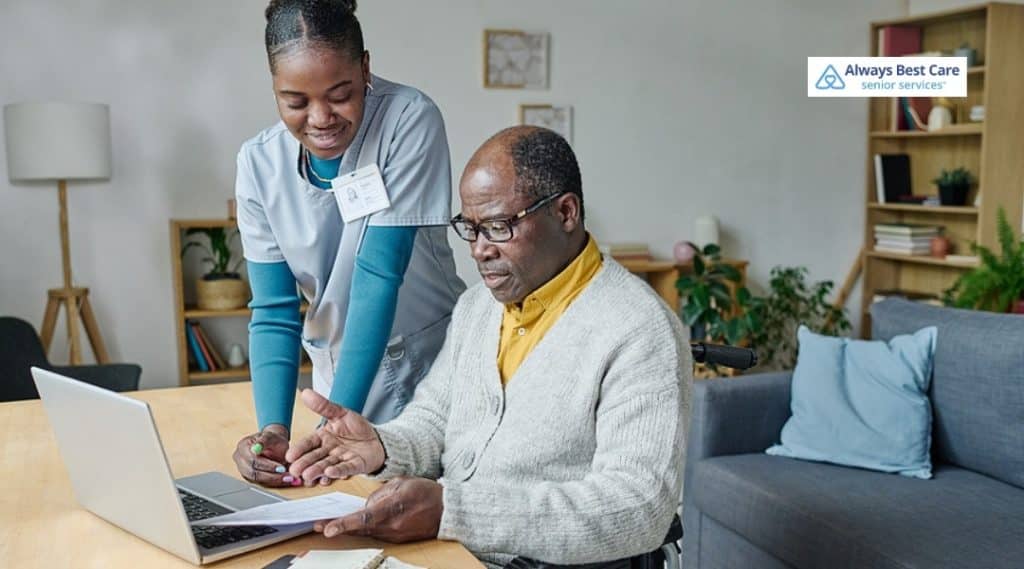 4. Caregiver in scrubs assists a senior in a wheelchair with documents beside a laptop at home (Always Best Care logo).