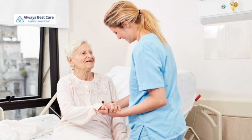 In-home caregiver helping an older adult put on a bright pink cardigan while they sit on the edge of the bed
