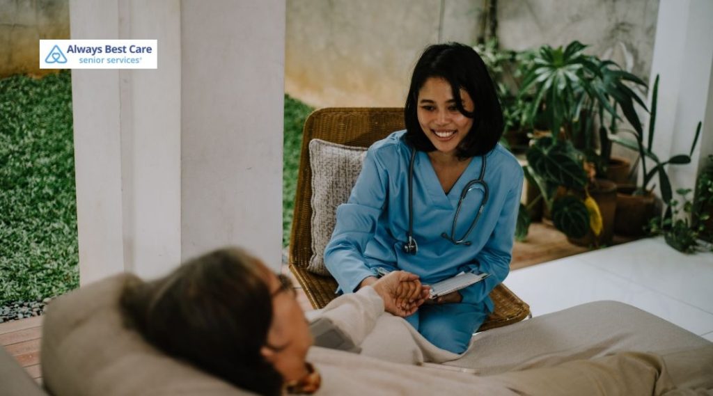 Caregiver in blue scrubs holding an older adult’s hand at the bedside in a medical setting, providing reassurance and support