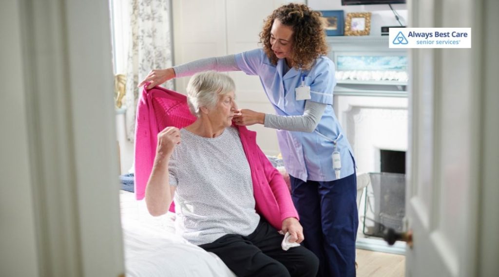 In-home caregiver helping an older adult put on a bright pink cardigan while they sit on the edge of the bed