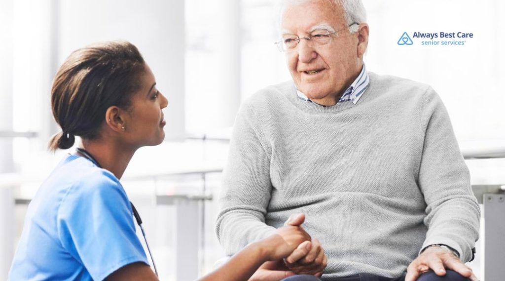 Nurse in blue scrubs sitting outdoors holding an older adult’s hand, offering comfort during a home visit.