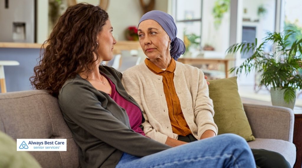 Two women sitting close together on a sofa, one wearing a purple headscarf, sharing a quiet conversation.