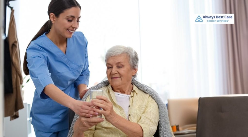 A caregiver helping an elderly woman with a cup of medication while both smile. Always Best Care logo in the bottom right corner.