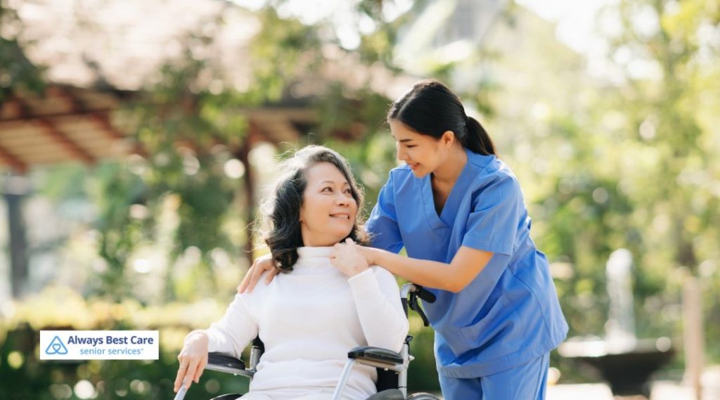 A caregiver assisting a senior woman in a wheelchair, helping her enjoy a walk outdoors in a garden-like setting. The caregiver provides both physical and emotional support, showing a caring bond.