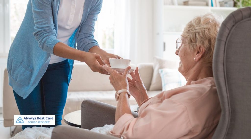 A caregiver offering a warm beverage to an elderly woman sitting comfortably in her living room. The caregiver smiles as the senior receives the drink, emphasizing thoughtful, personalized home care.