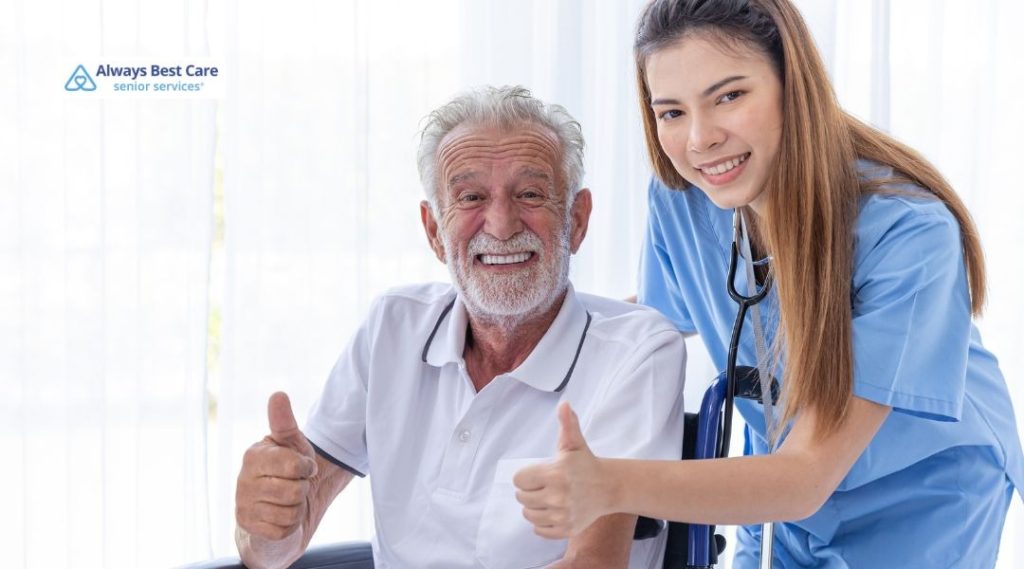 An elderly man and a caregiver both give a thumbs up, smiling happily. The caregiver, wearing a blue uniform, is helping the senior in a wheelchair, showing a strong bond and positive energy.