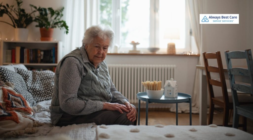 2. Older woman sitting on the edge of her bed in a cozy room, looking thoughtful near a small side table (Always Best Care logo).