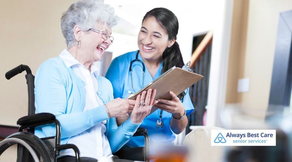 A caregiver from Always Best Care of Thousand Oaks smiles and interacts with a senior woman in a wheelchair, reviewing care notes in a friendly, caring environment.