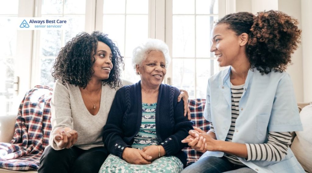 A caregiver, a senior woman, and a family member sit together on a couch, laughing and engaging in conversation. The caregiver offers a supportive and friendly presence, ensuring the senior woman feels comfortable.