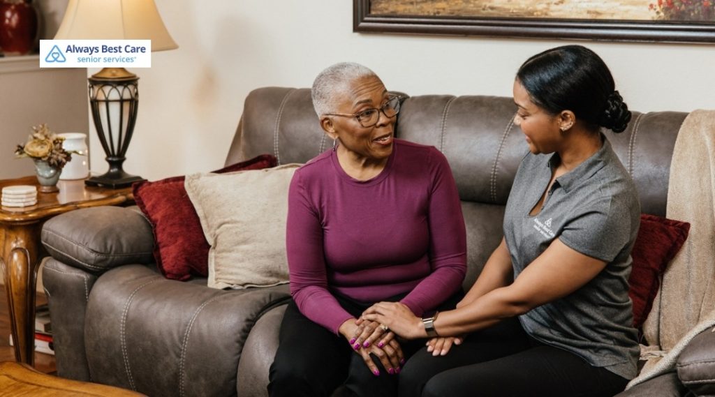 Caregiver holding senior woman’s hands while talking on living room couch