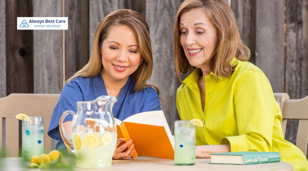 Caregiver reading a book with a senior woman while sharing lemonade outdoors