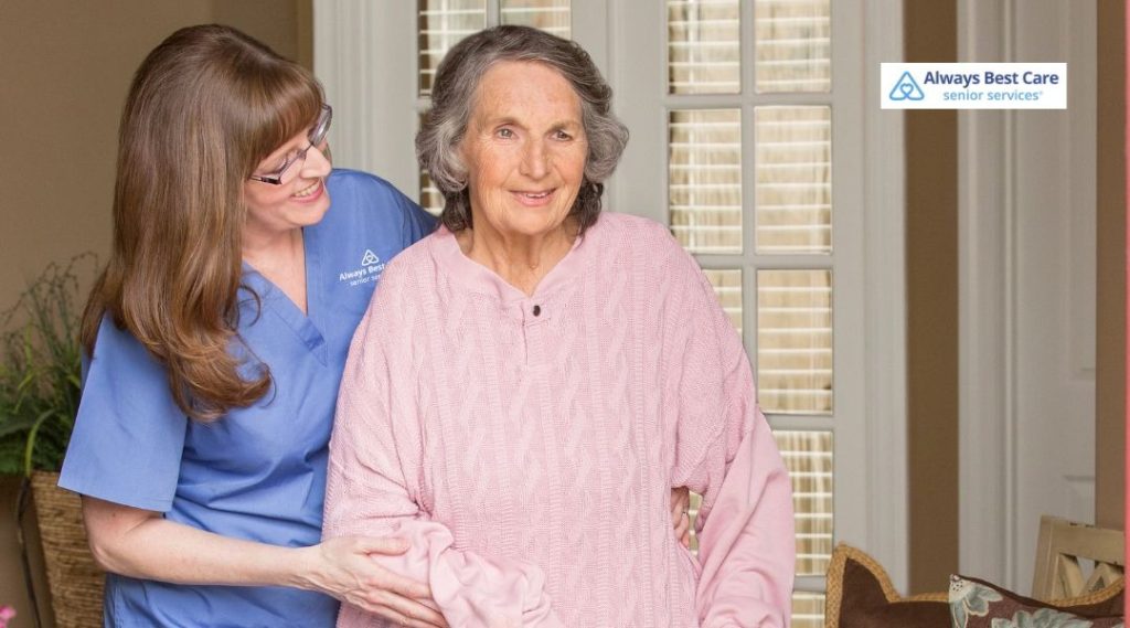 In-home caregiver in blue scrubs gently supporting an older woman as she stands up in the living room.