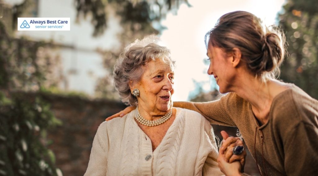 Younger woman leaning toward a well-dressed older woman outdoors, holding her hand and offering support, Always Best Care logo in corner.
