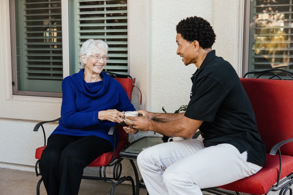 A caregiver hands a cup of tea to an elderly woman seated on a porch.