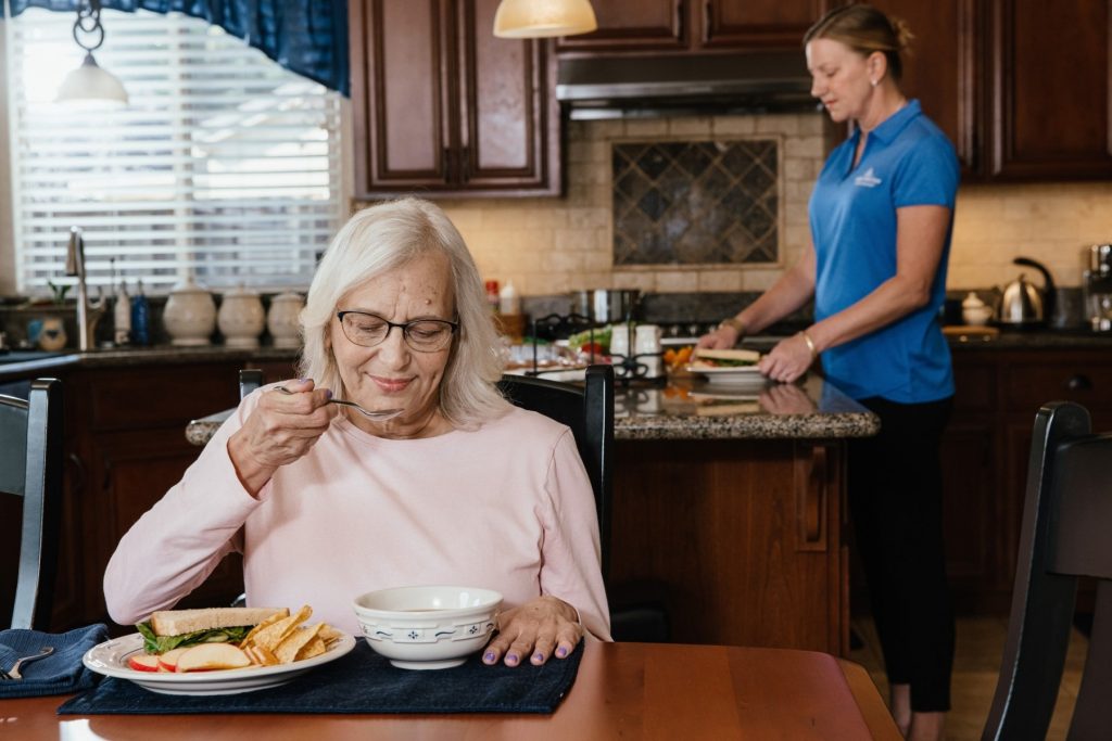 An older woman eats at a kitchen table while a caregiver prepares food in the background.