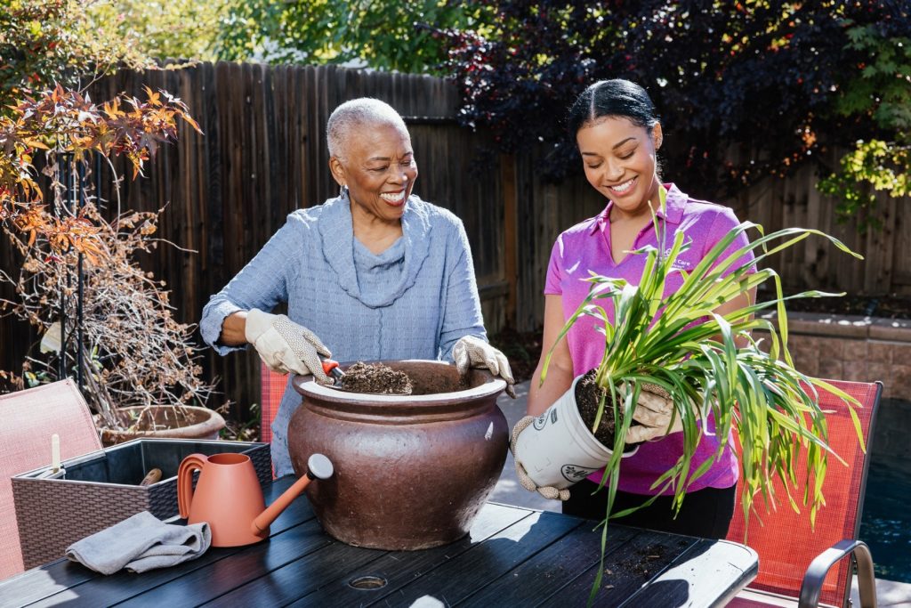 Two caregivers smile while potting a plant outdoors at a backyard table.
