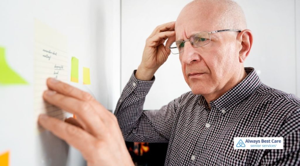 Older man wearing glasses standing in front of a board, concentrating on handwritten reminder notes and sticky notes, appearing to focus carefully, with the Always Best Care senior services logo in the corner.