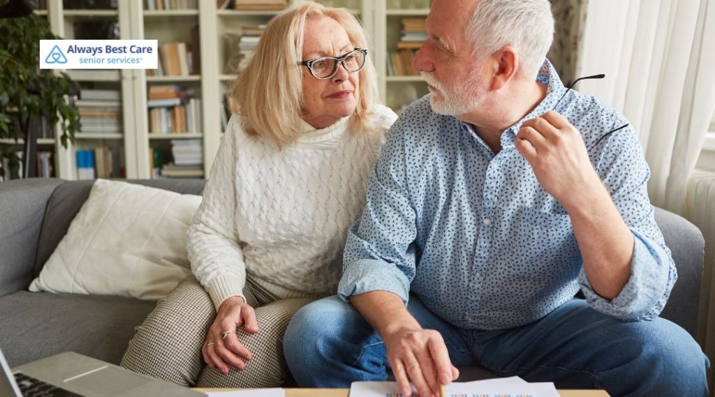 Older couple sitting on a couch, looking at each other while reviewing documents and charts on the table, suggesting a serious conversation about plans or finances, with the Always Best Care senior services logo in the corner.