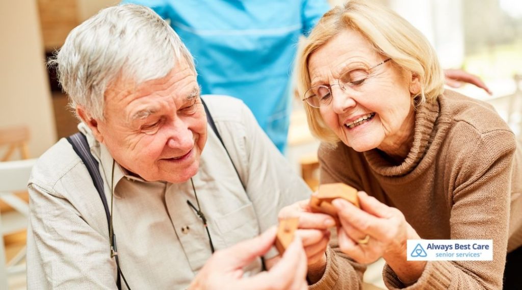 Smiling older couple sitting together at a table, happily working with small wooden blocks, with a caregiver in the background and the Always Best Care senior services logo in the corner.