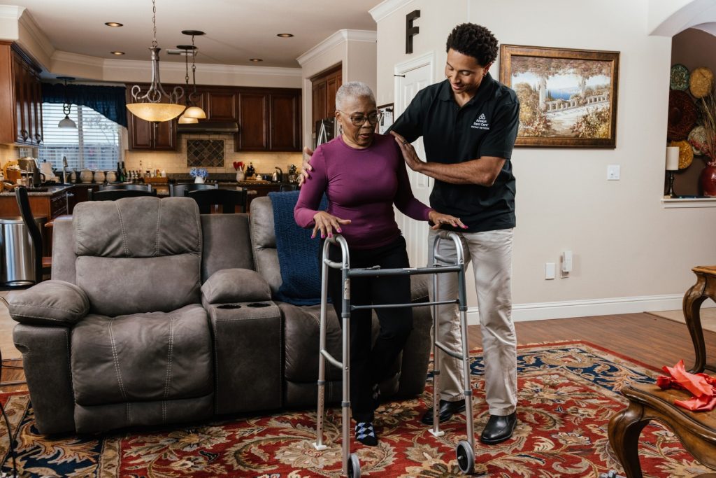 Caregiver assists an older woman as she stands and uses a walker in her home.