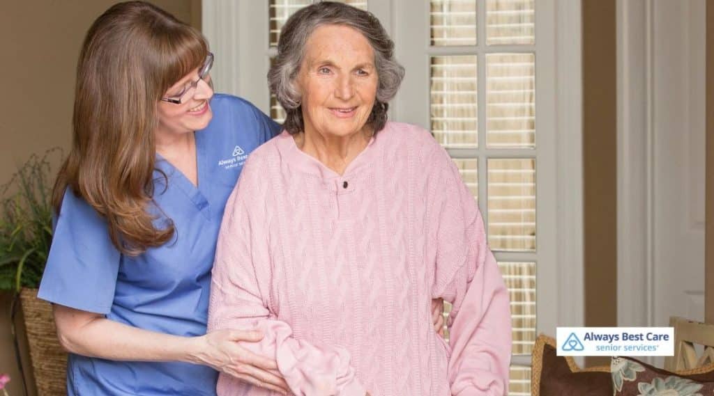 A caregiver helps an elderly woman put on her sweater, both smiling and engaged in a caring, supportive moment. The background features a bright and inviting home environment.