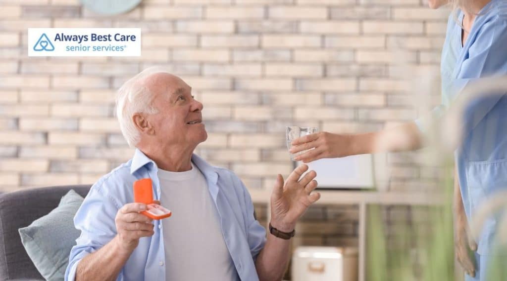 An elderly man smiles as a caregiver hands him a glass of water while he holds his medication box. They share a friendly moment in a bright, welcoming room.