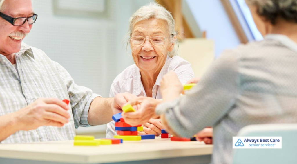 Group of smiling seniors sitting at a table, stacking colorful blocks together during a memory care activity, Always Best Care senior services logo in the corner.