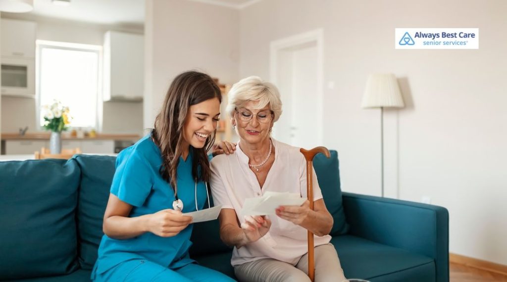 Caregiver in scrubs sitting on a sofa with a senior woman who uses a cane, looking through old photographs together in a cozy home, Always Best Care senior services logo in the corner.