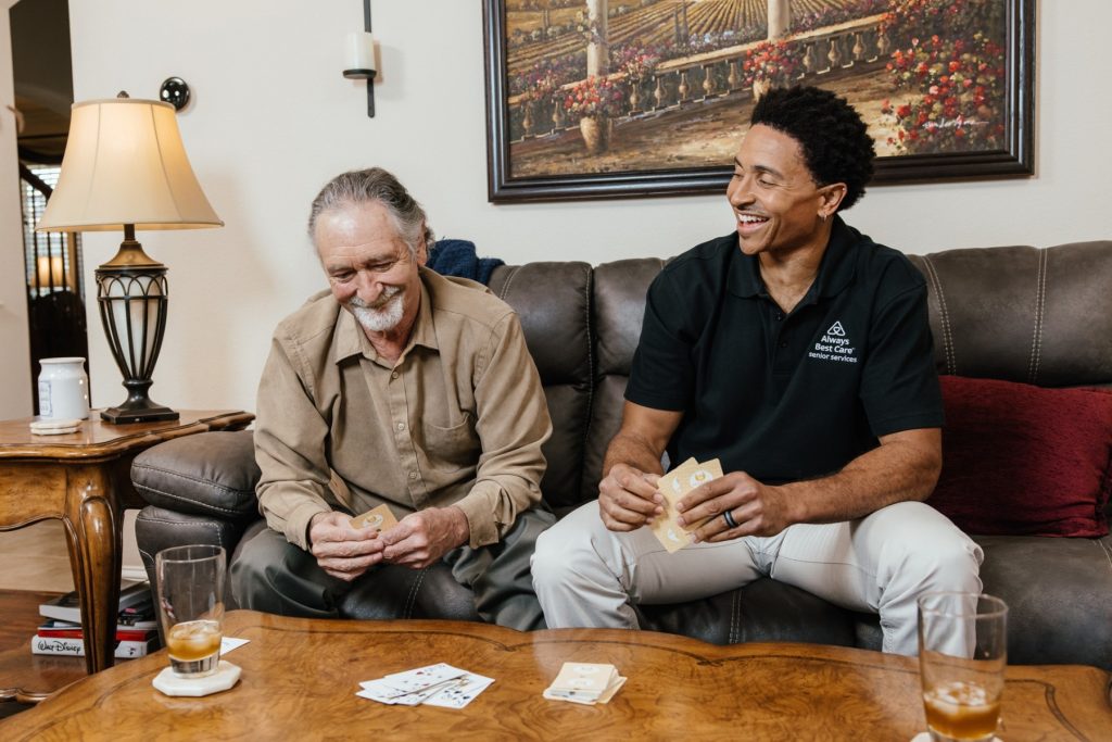 Male caregiver in a black polo shirt plays cards on a coffee table with an older man, both laughing while sitting on a living room couch