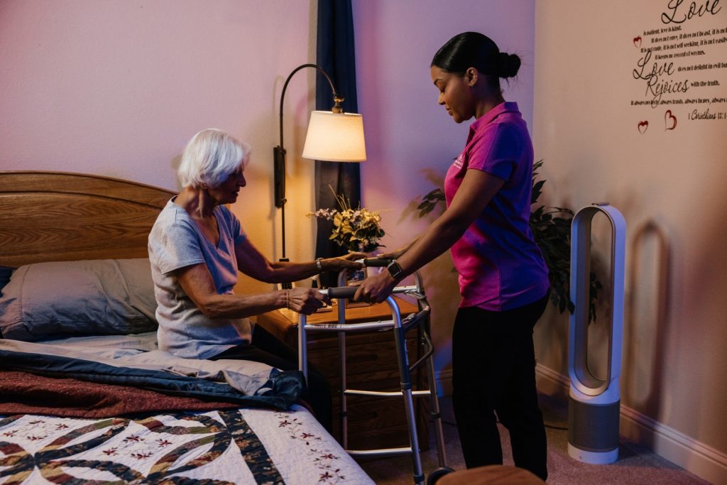 Smiling caregiver in a pink polo shirt sits on a sofa with an older woman, and they look through a photo album together in a bright living room.