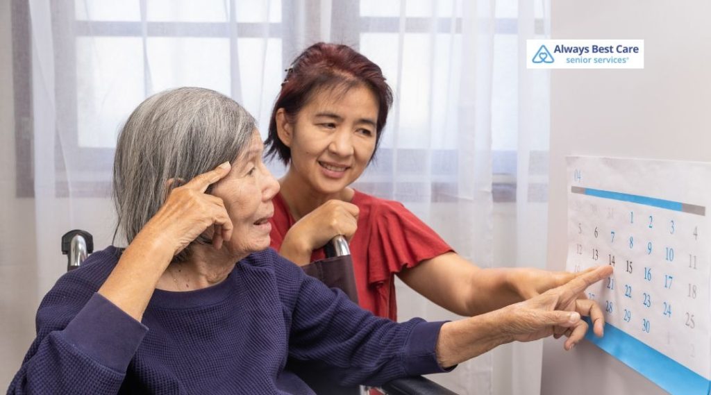 Caregiver gently guiding an older woman in a wheelchair as they look together at a wall calendar, discussing upcoming dates.