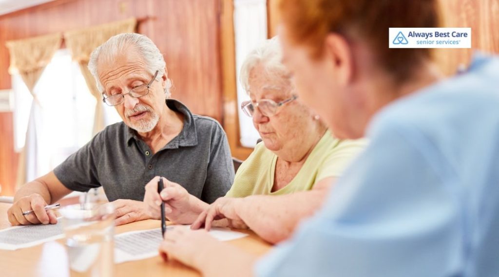 Senior couple reviewing paperwork at a table with a caregiver, who points to a document as they discuss important information.
