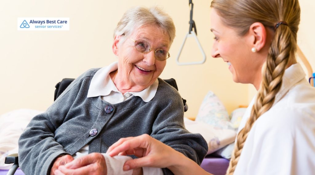 A caregiver is assisting a joyful elderly woman in a wheelchair, helping her with her clothing. Both women are smiling, and the scene conveys warmth and trust.
