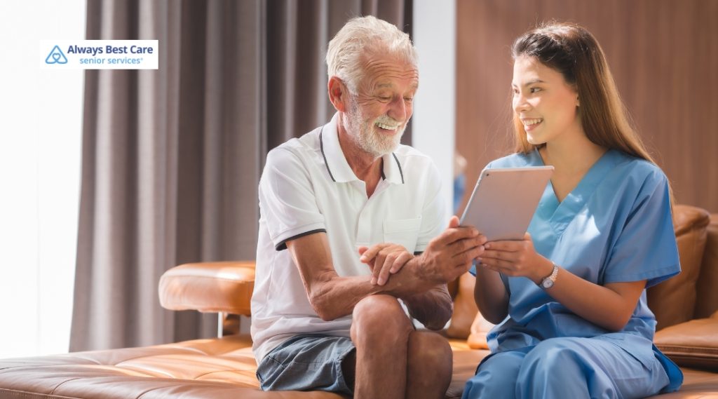 A caregiver and an elderly man share a moment of joy as they look at a tablet together. The elderly man smiles brightly, and the caregiver, in scrubs, appears engaged and friendly.